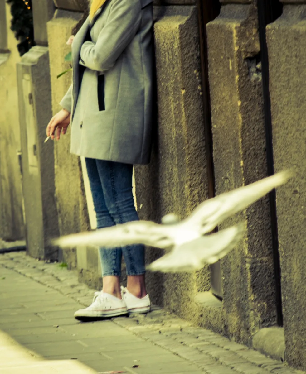 Woman leaning against a stone wall with a cigarette as a white bird flies past